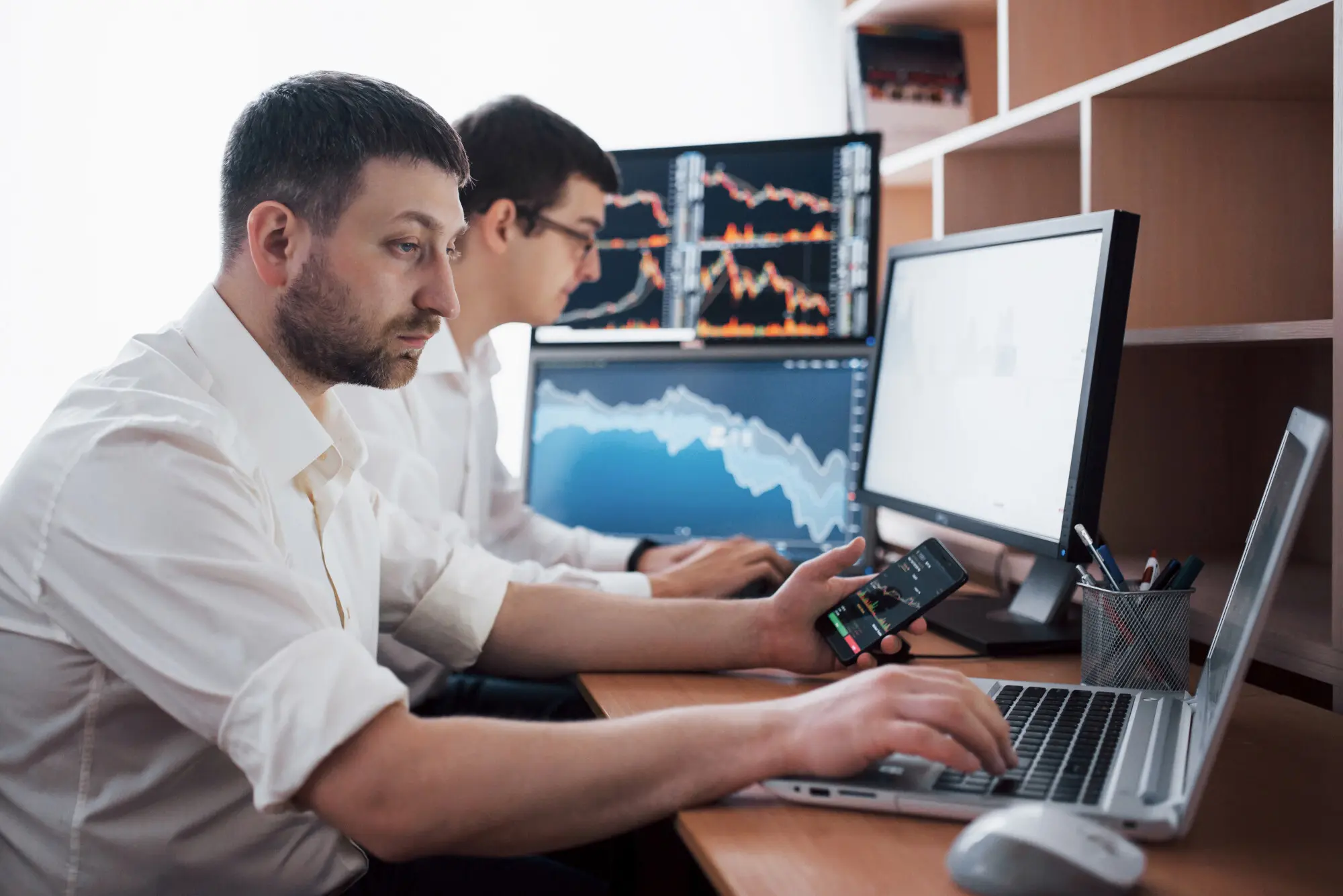 Ein Team von Börsenmaklern führt in einem dunklen Büro mit Bildschirmen ein Gespräch. Sie analysieren Daten, Diagramme sowie Berichte für Investitionszwecke. Kreative Teamarbeit der Trader, ohne den praktischen Nutzen aus dem Blick zu verlieren.